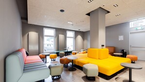 Espaço lounge moderno com assentos, mesas e sinalização indicando amenidades como salas de conferência, cozinha para hóspedes e armários.