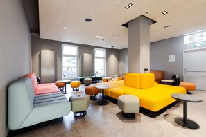 Un espacio lounge moderno con asientos, mesas y un letrero que indica servicios como salas de reuniones, cocina para huéspedes y taquillas.