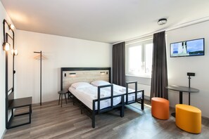 Una habitación de hotel moderna con una cama, una mesita, dos otomanas, un televisor y un ventanal con cortinas.