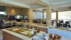 Buffet com diversas opções de comida, área de refeições com mesas e cadeiras, e uma grande janela que oferece luz natural.