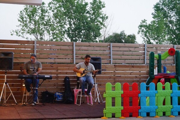 Two musicians performing on a stage with a wooden fence backdrop and colorful playground equipment in the foreground. - Selyria Resort (Zakynthos)