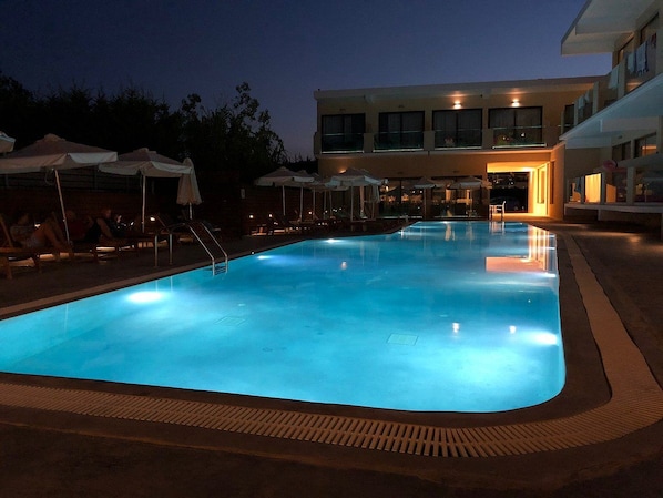 Ein Schwimmbecken mit Liegestühlen und Sonnenschirmen, umgeben von einem Gebäude mit mehreren Stockwerken und Fenstern.