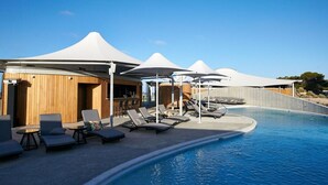 Zona junto a la piscina con sillones de descanso y sombrillas, una estructura de madera y un cielo azul despejado.
