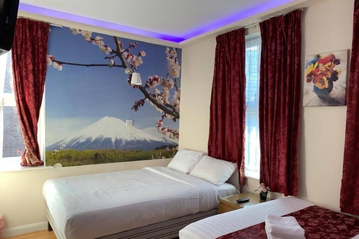 Ein Hotelzimmer mit zwei Betten, einem Wandgemälde eines Berges, roten Vorhängen und einem Nachttisch mit einer Lampe.