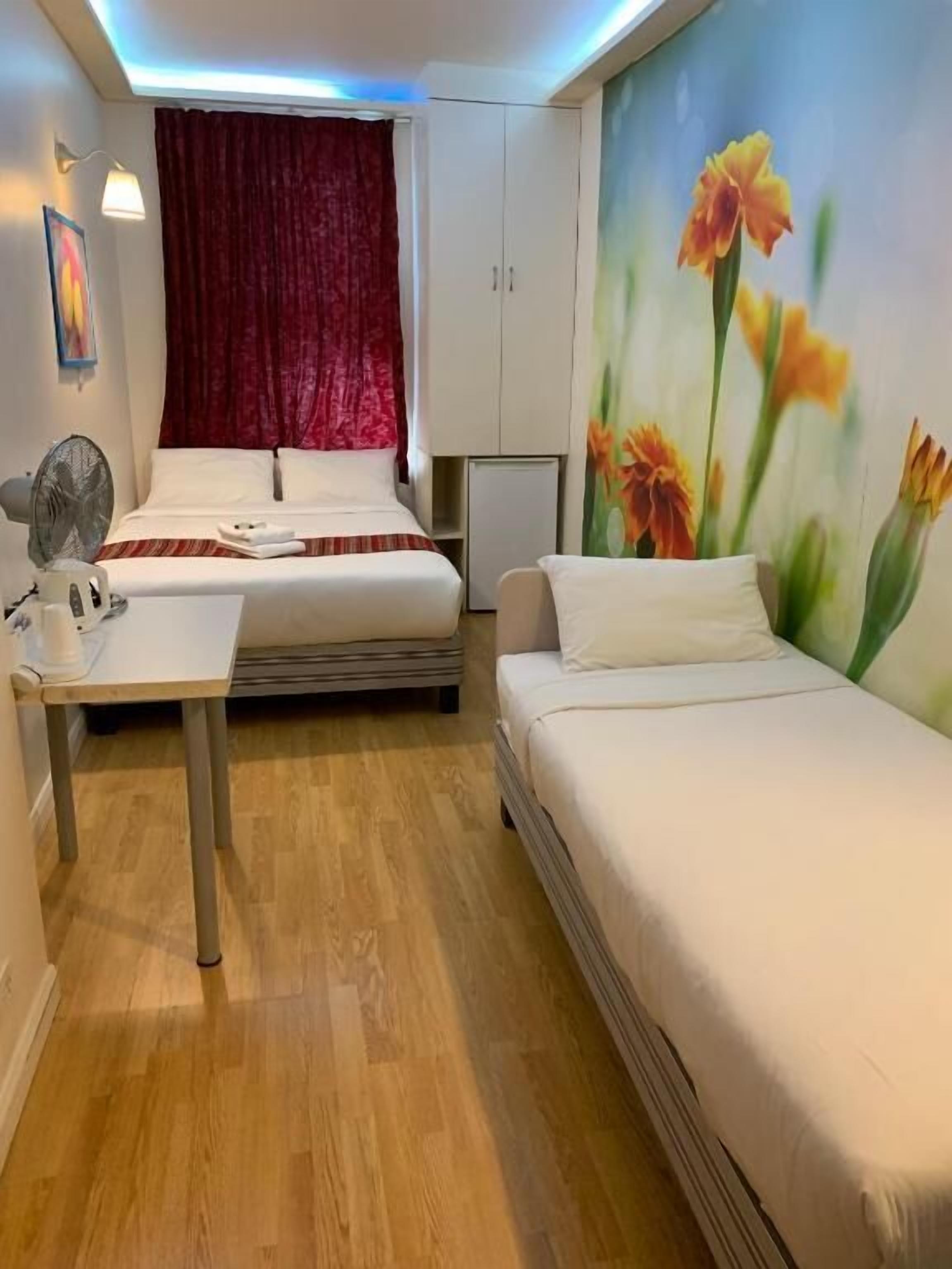 Habitación de hotel con dos camas, un escritorio, un ventilador, un refrigerador y una pared con un mural floral.