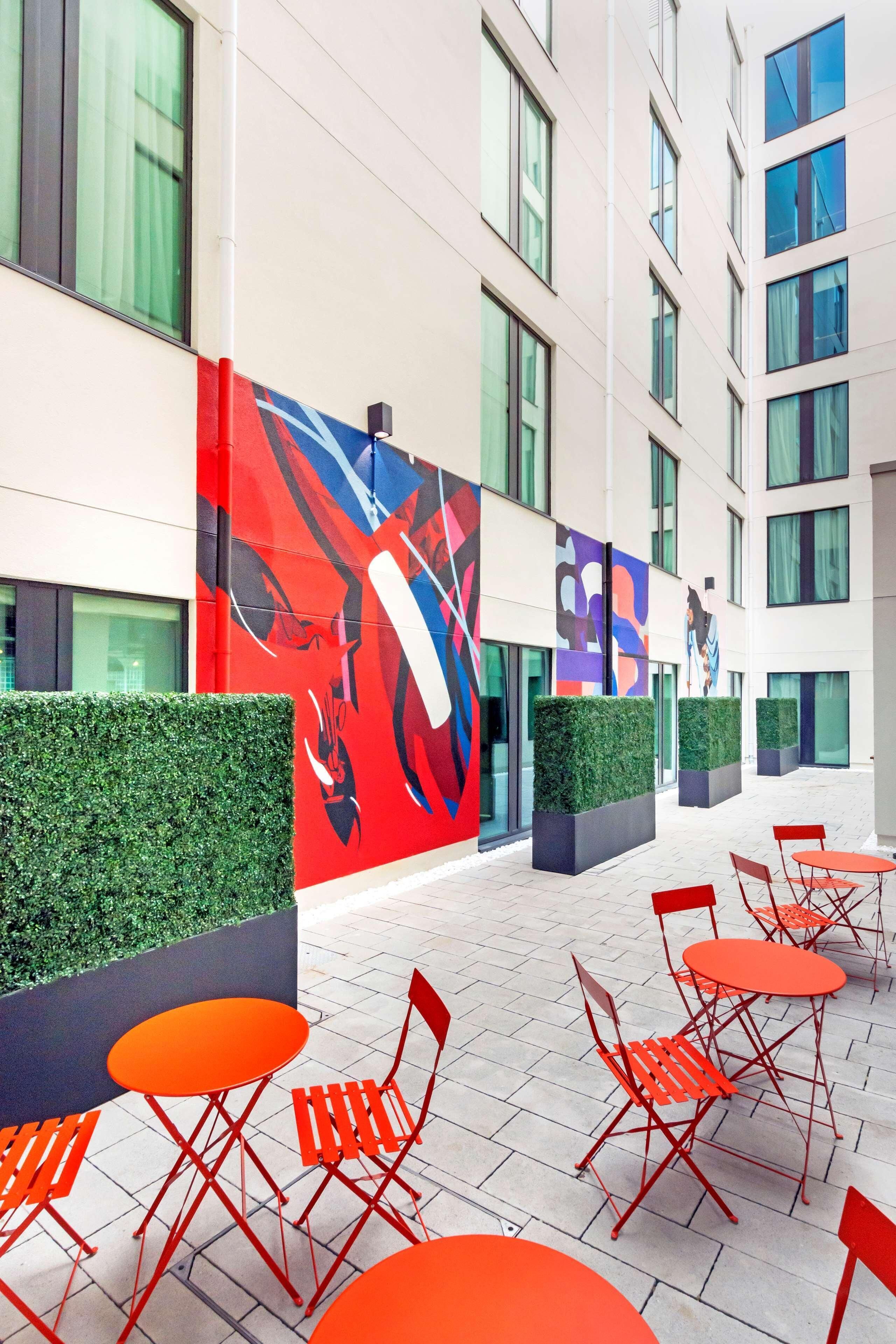 A modern outdoor seating area with red chairs and tables, a large abstract mural, and a building with multiple windows and a green hedge.