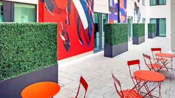 A modern outdoor seating area with red chairs and tables, a large abstract mural, and a building with multiple windows and a green hedge.