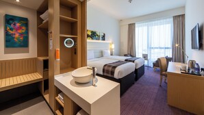 A hotel room with a large bed, a desk, a chair, a round mirror, a sink, and artwork on the wall. - Premier Inn Dragon Mart Hotel (Dubai)