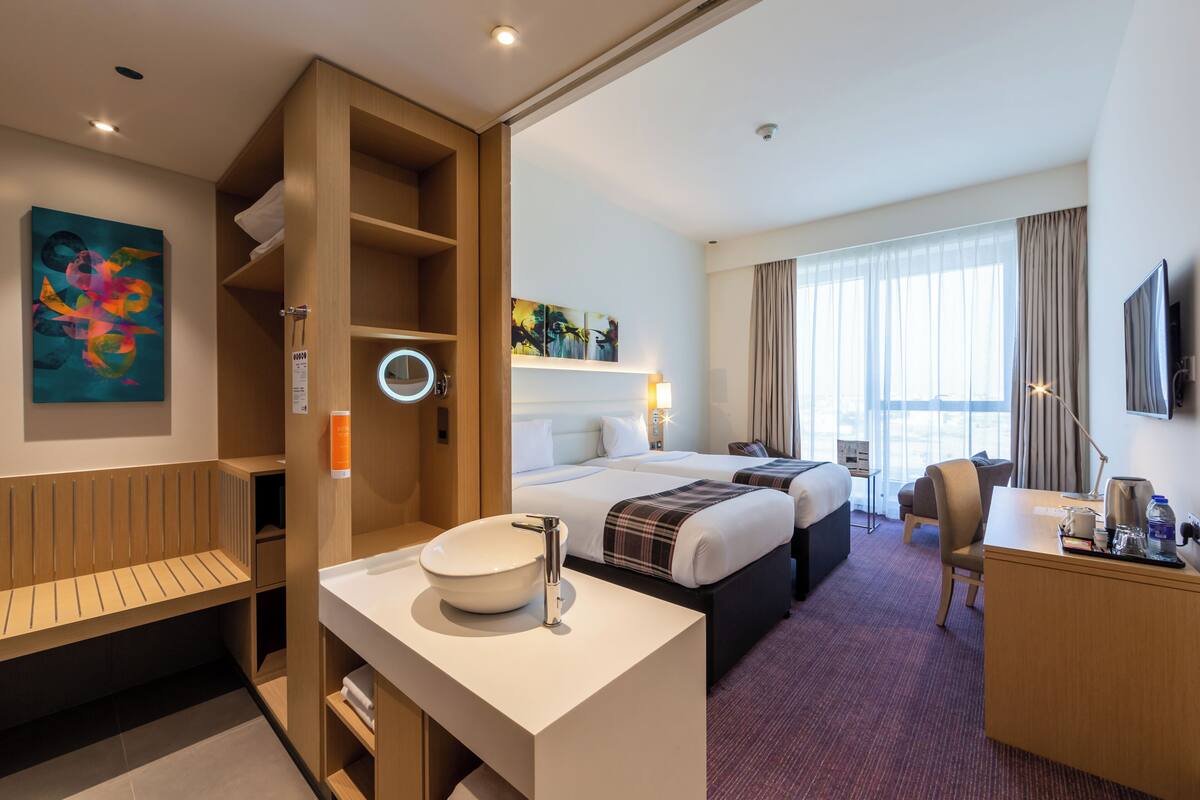 Hotellihuone, jossa on suuri sänky, työpöytä, tuoli, pyöreä peili, pesuallas ja taideteoksia seinällä.