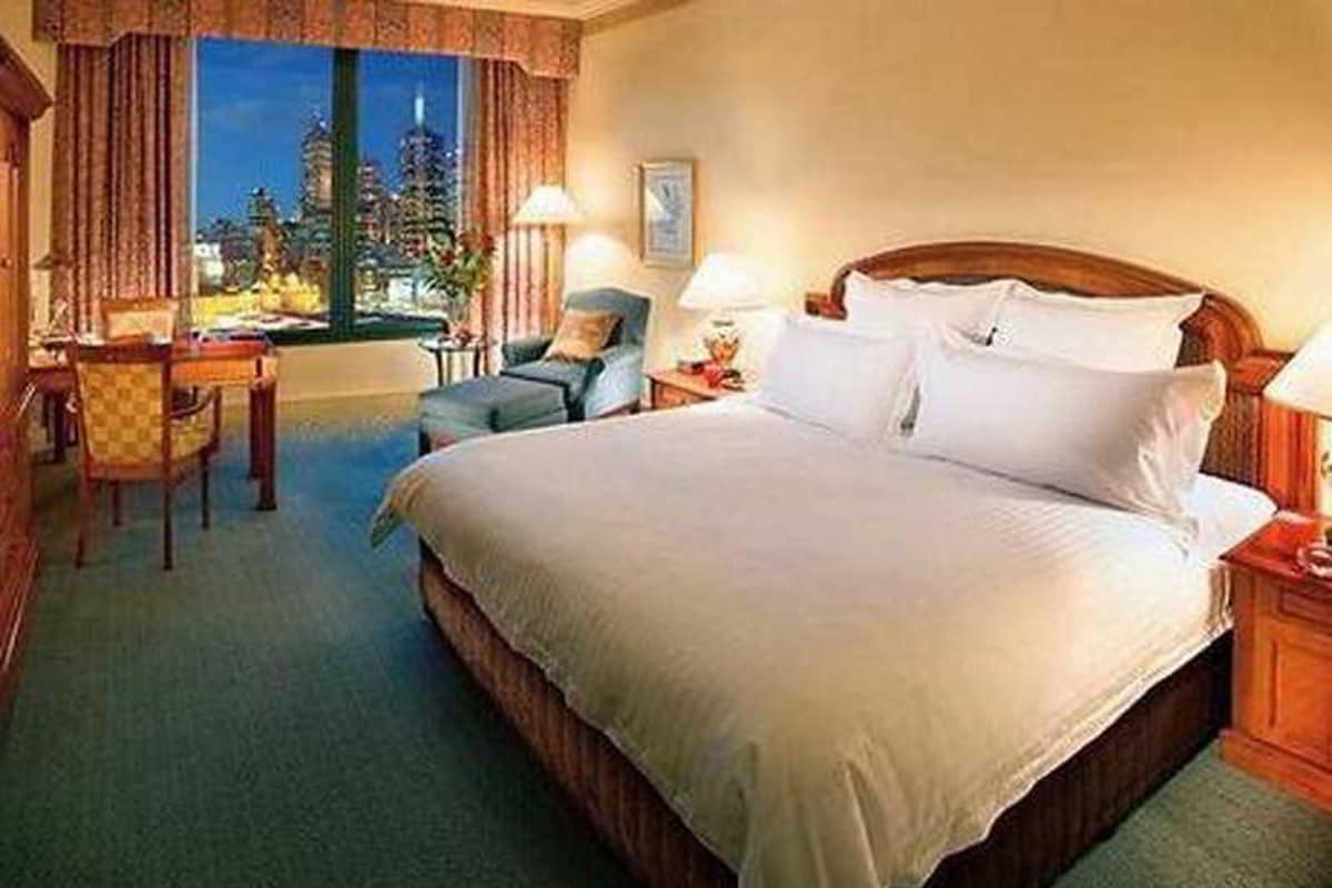 A hotel room with a large bed, a desk, and a chair. There is a window with a view of a cityscape.