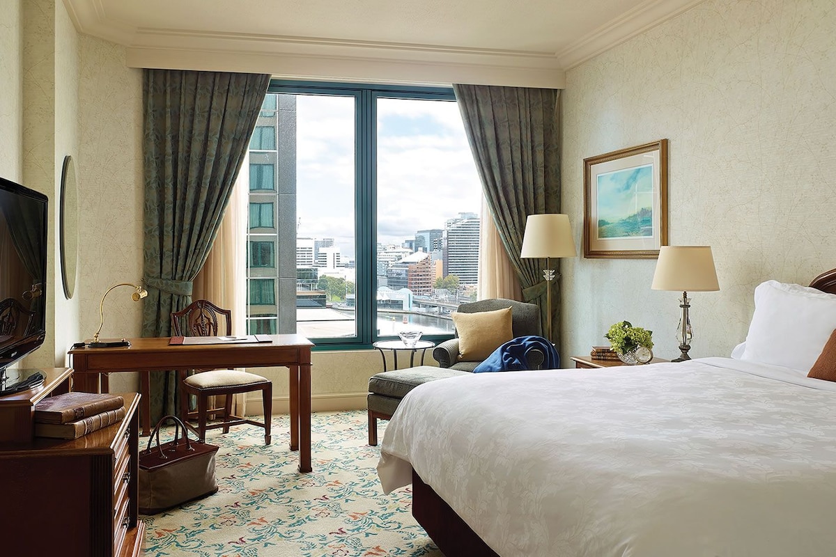 A hotel room with a large bed, a desk with a chair, a television, and a view of the cityscape through the window.