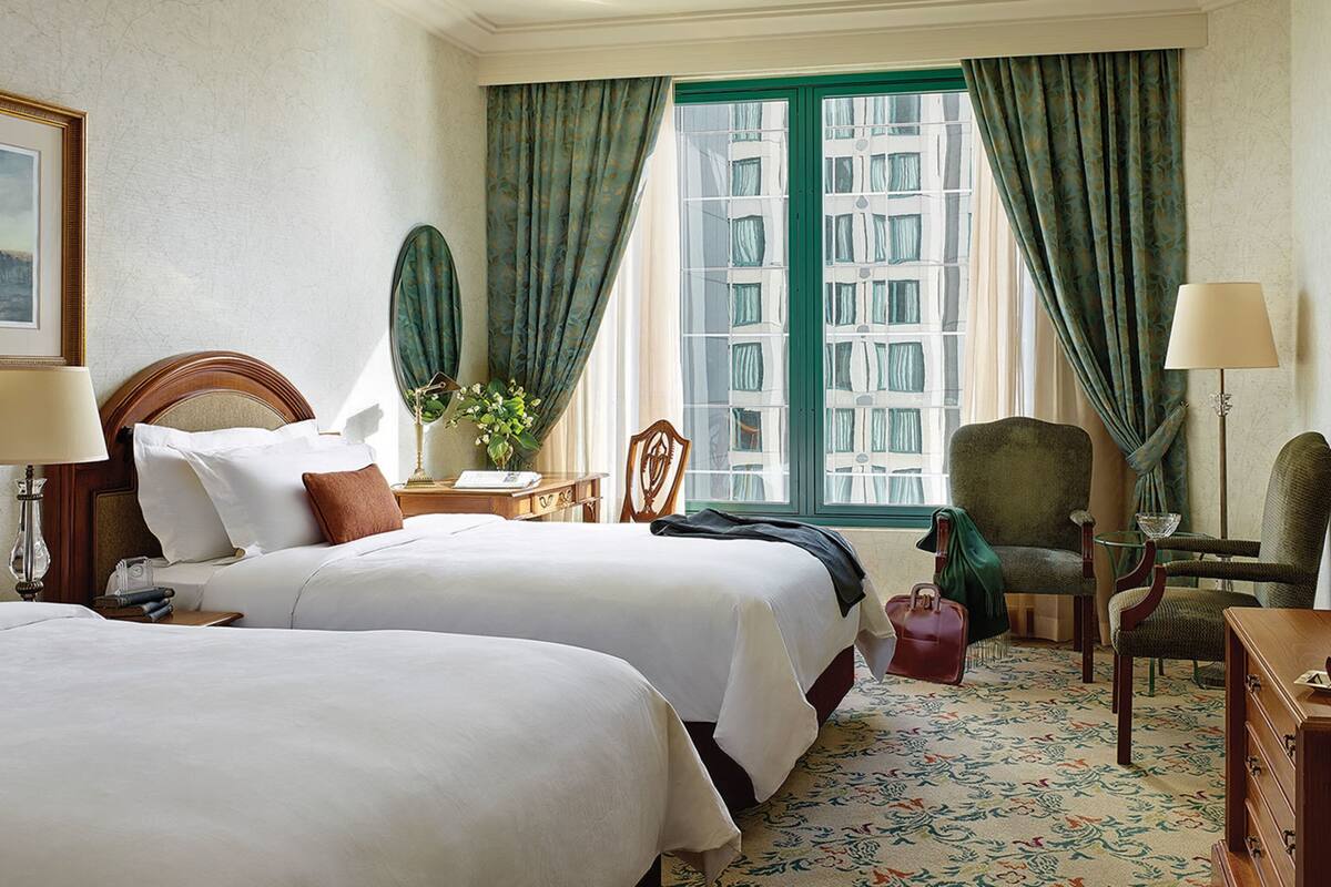 A hotel room with two beds, a television, a desk with a lamp, a chair, and a view of buildings through the window.