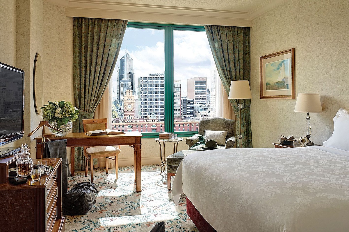 A hotel room with a bed, desk, chair, and a view of city buildings.