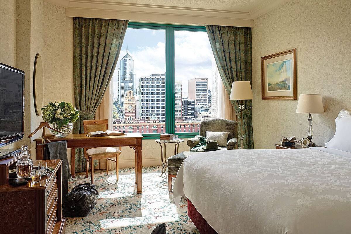 A hotel room with a bed, desk, chair, and a view of city buildings.