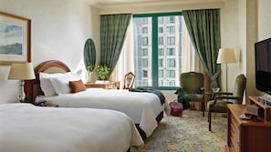 Ein Hotelzimmer mit zwei Betten, einem Fernseher, einem Schreibtisch mit Lampe, einem Sessel und Blick auf die Stadt durch große Fenster.