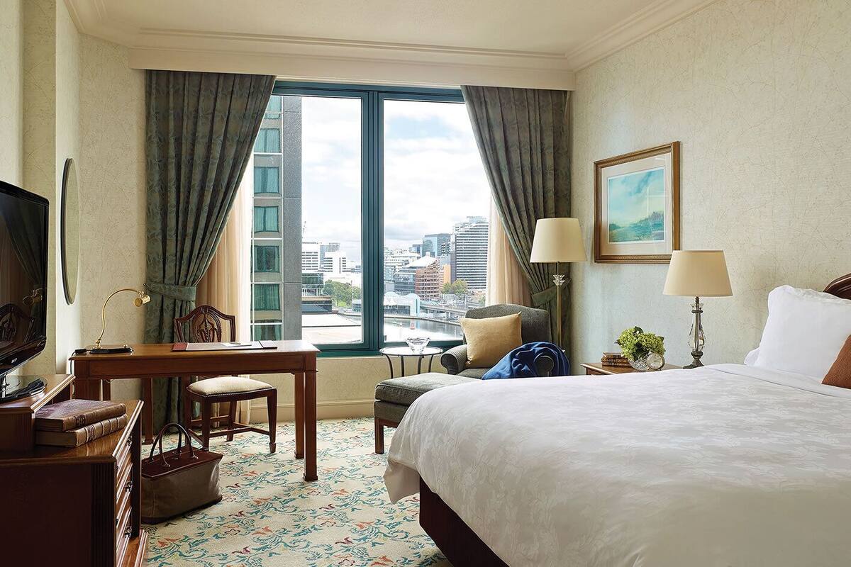 A hotel room with a large bed, a desk with a chair, a television, and a view of the cityscape through the window.