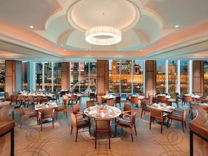 Ein geräumiger Essbereich mit mehreren Tischen und Stühlen, große Fenster mit Blick auf die Stadt und ein zentraler Kronleuchter.