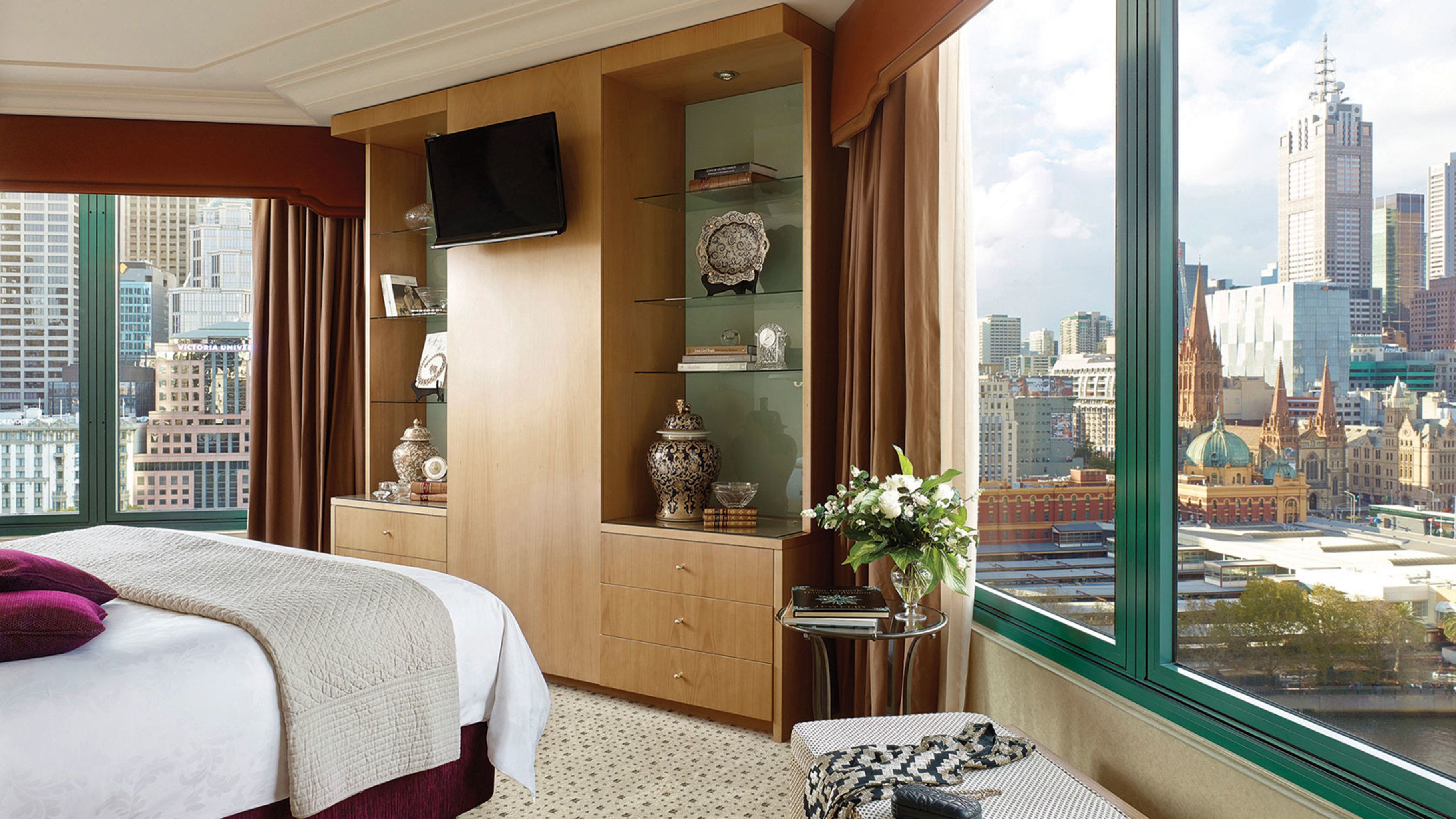 A hotel room with a large bed, a television mounted on the wall, a small table with a vase of flowers, and a view of a cityscape through large windows.
