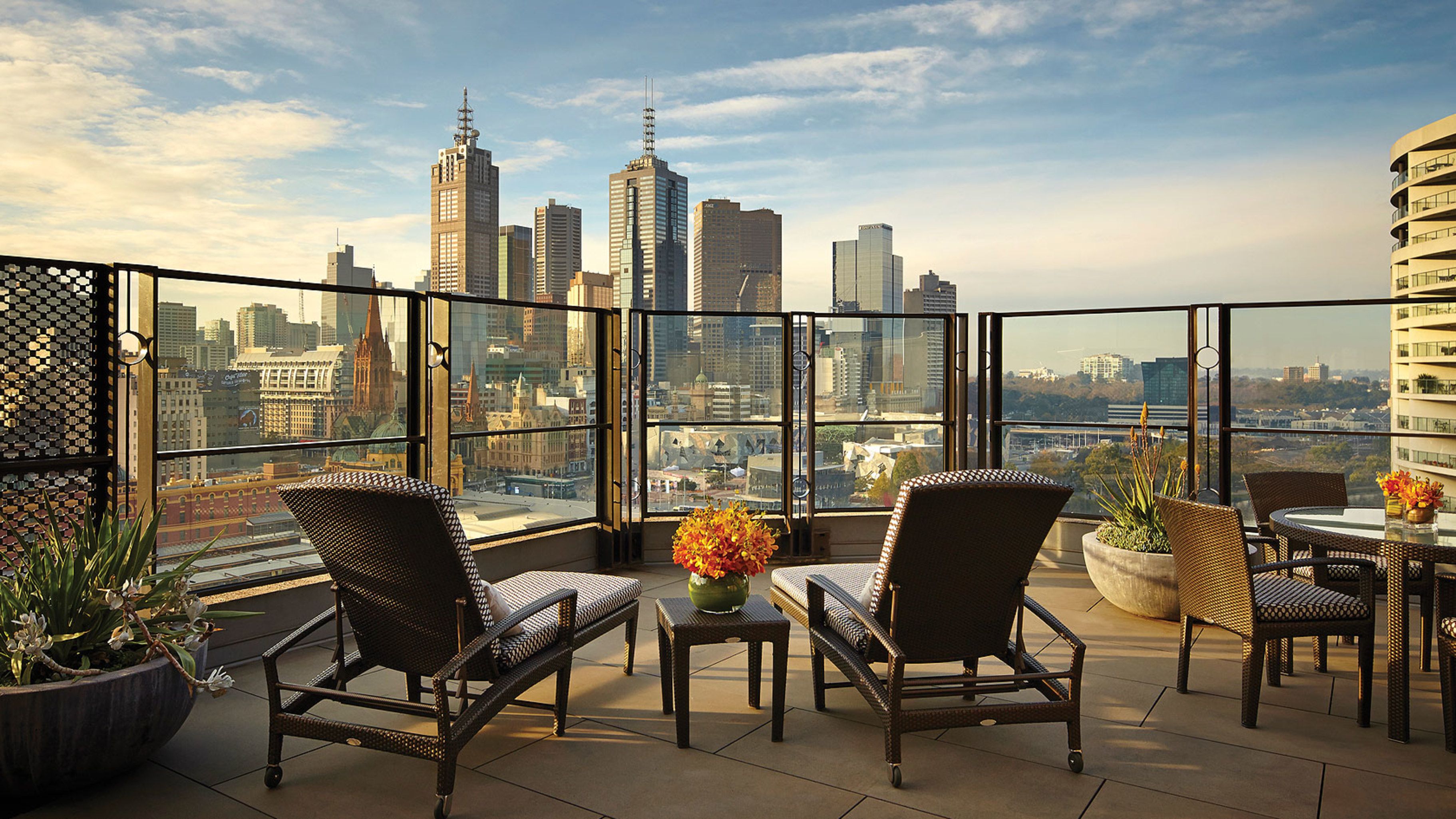 A terrace with wicker chairs, a table, and potted plants, overlooking a cityscape with tall buildings.
