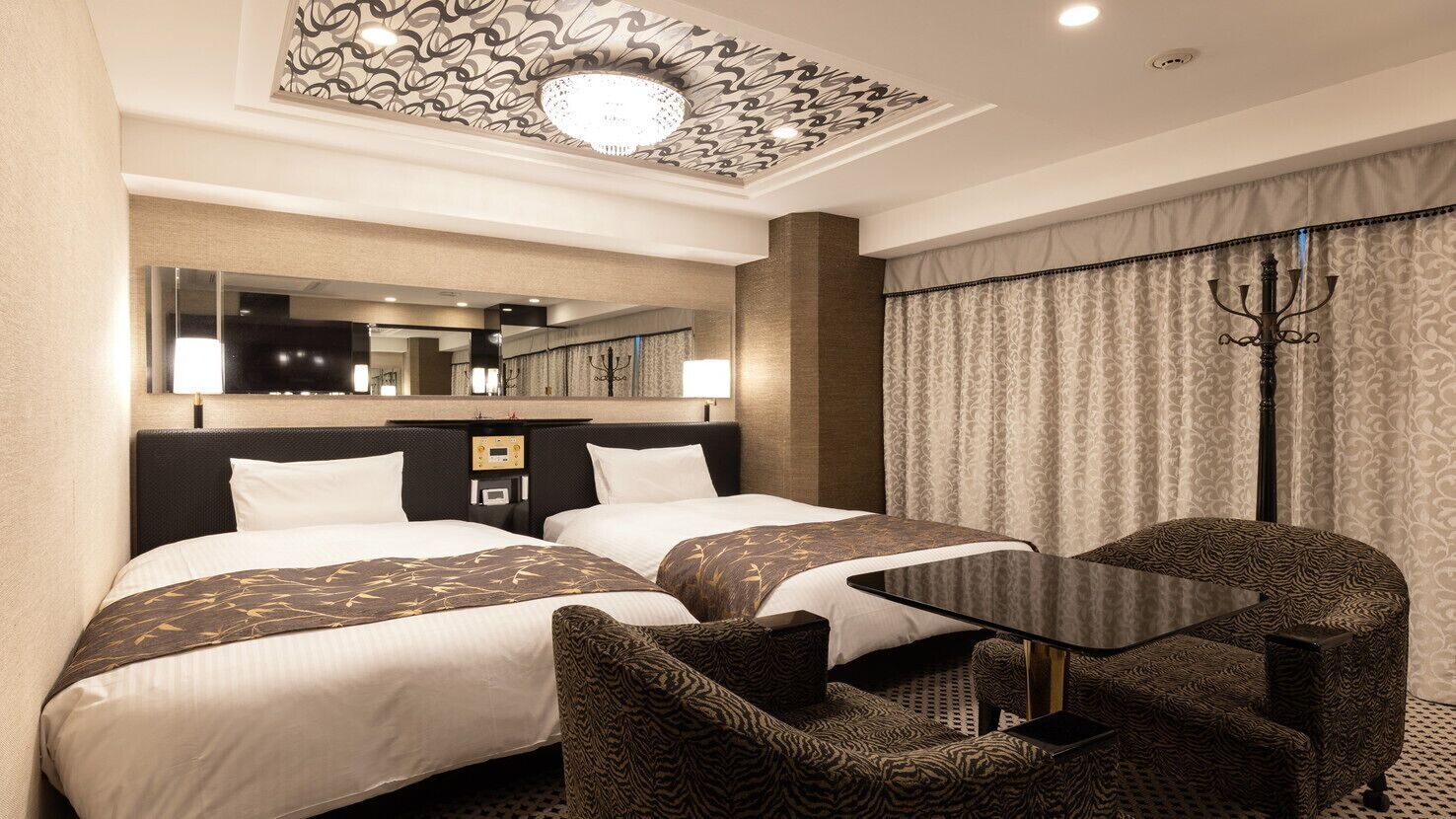 A hotel room with two beds, a desk, a chair, a mirror, and a chandelier.