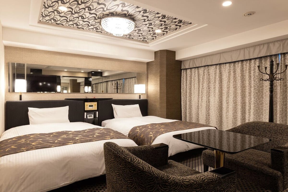 A hotel room with two beds, a desk, a chair, a mirror, and a chandelier.