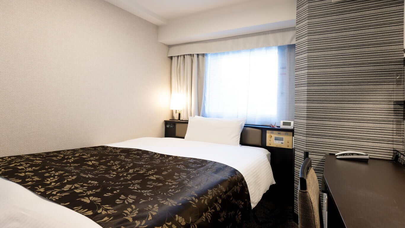 A hotel room with a bed, a bedside table, a lamp, a window with curtains, and a wall with a textured pattern.