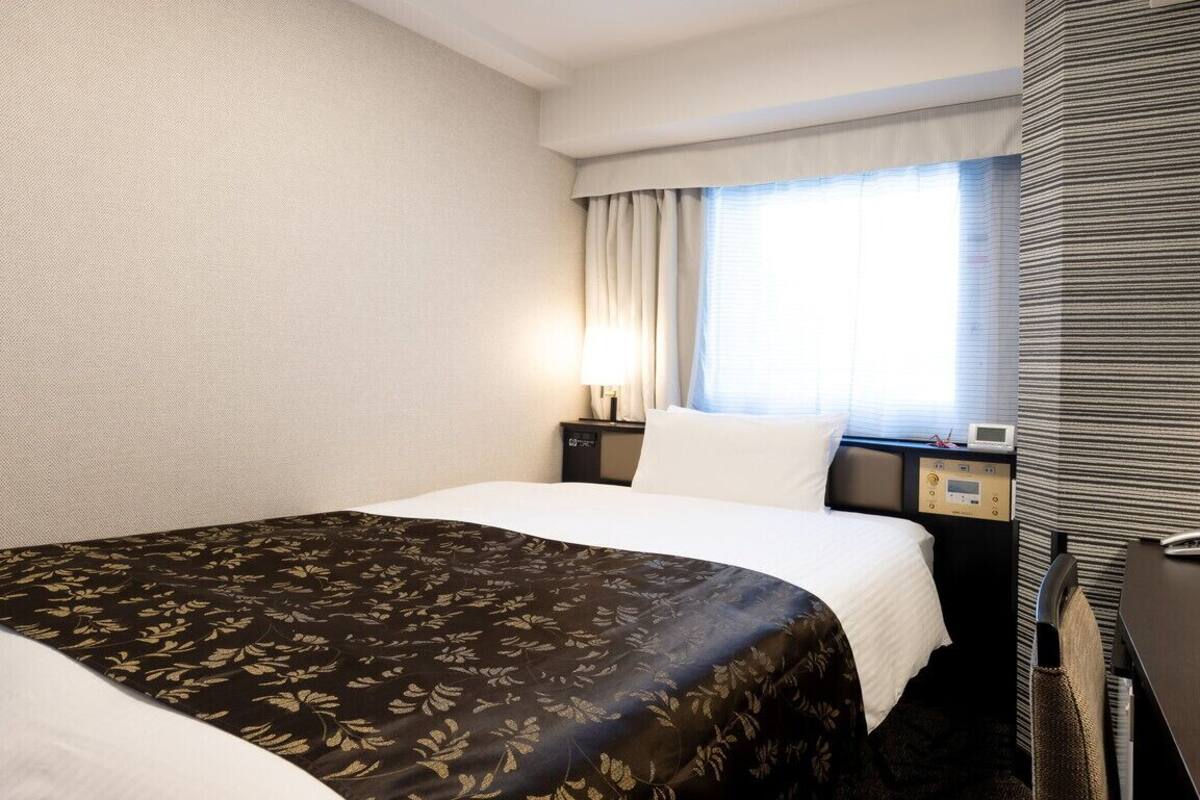 A hotel room with a bed, a bedside table, a lamp, a window with curtains, and a wall with a textured pattern.
