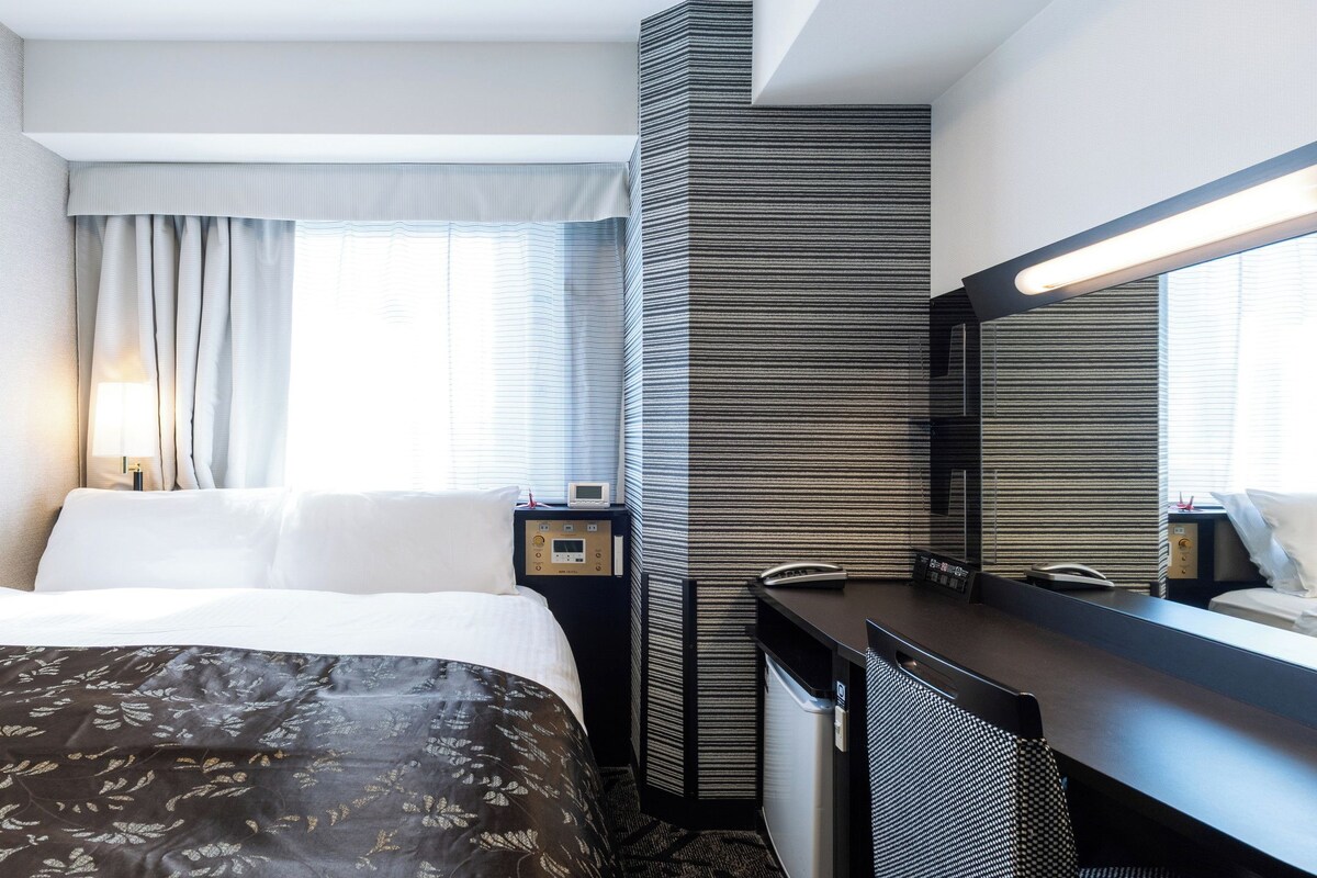 A hotel room with a bed, a desk, a mirror, and a window with curtains.