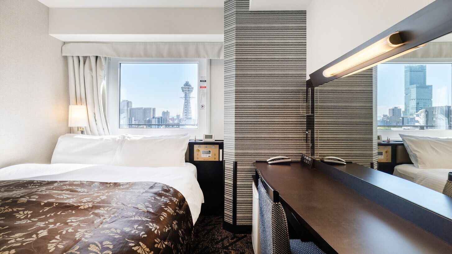 A hotel room with a bed, a desk, a mirror, a window with a city view, and a wall-mounted lamp.