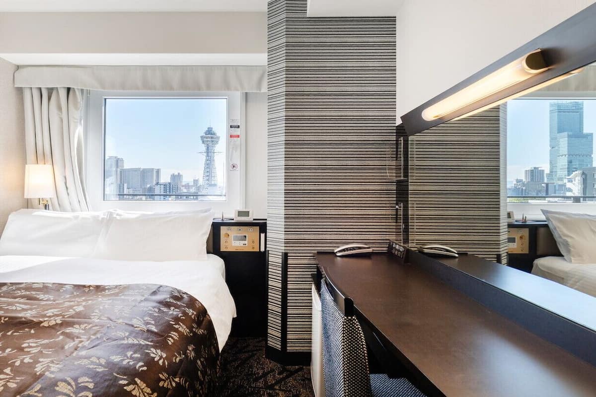 A hotel room with a bed, a desk, a mirror, a window with a city view, and a wall-mounted lamp.
