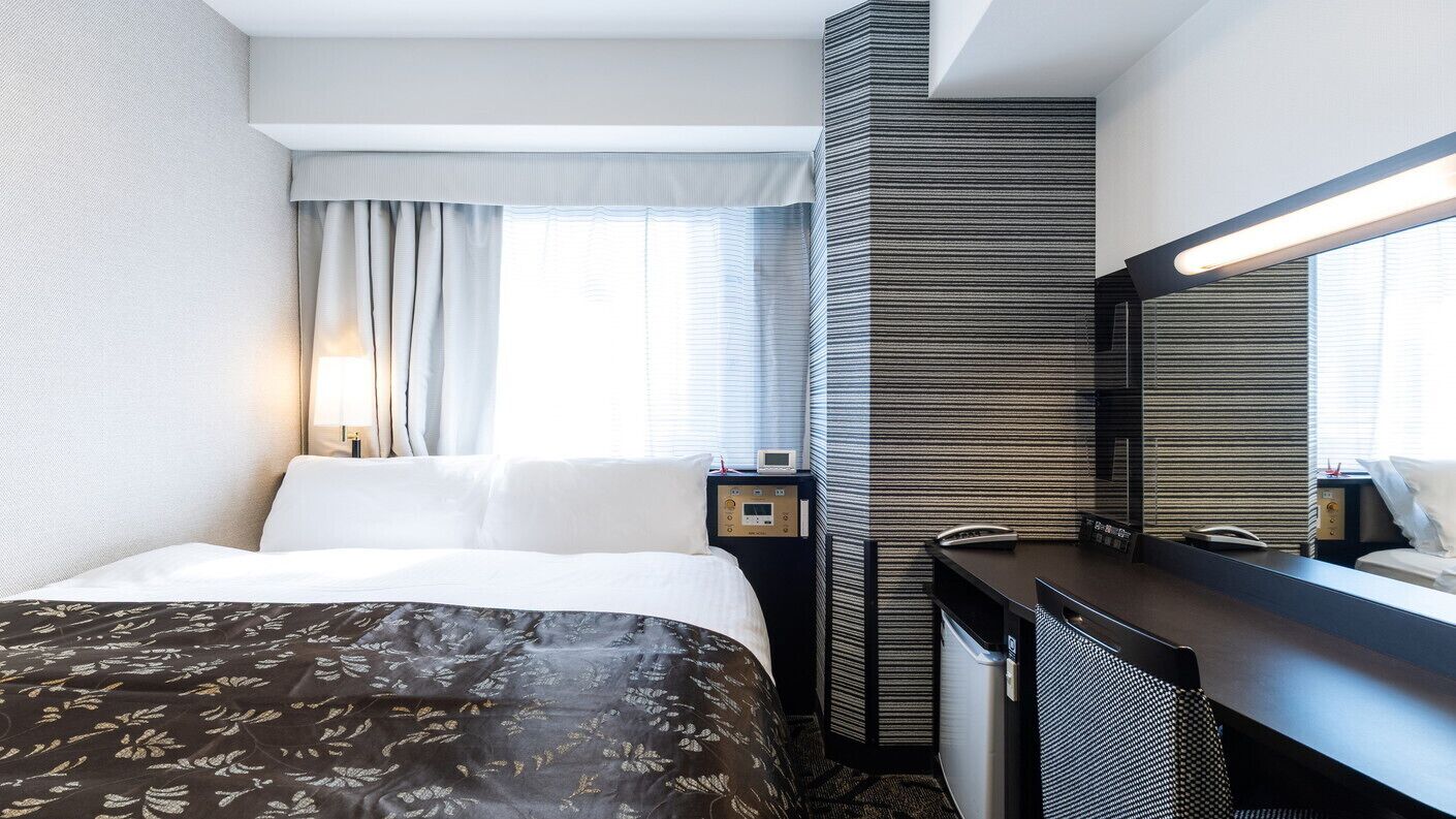 A hotel room with a bed, a desk, a mirror, a window with curtains, and a wall with a textured pattern.