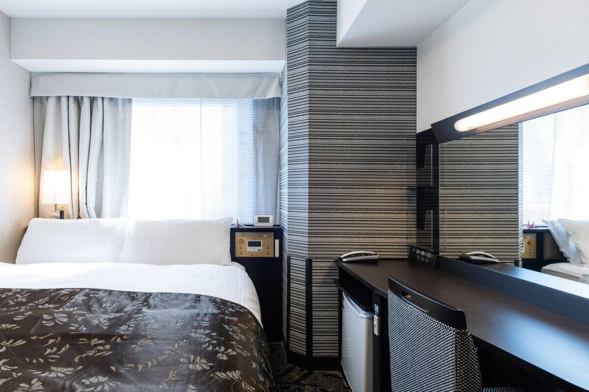 A hotel room with a bed, a desk, a mirror, a window with curtains, and a wall with a textured pattern.