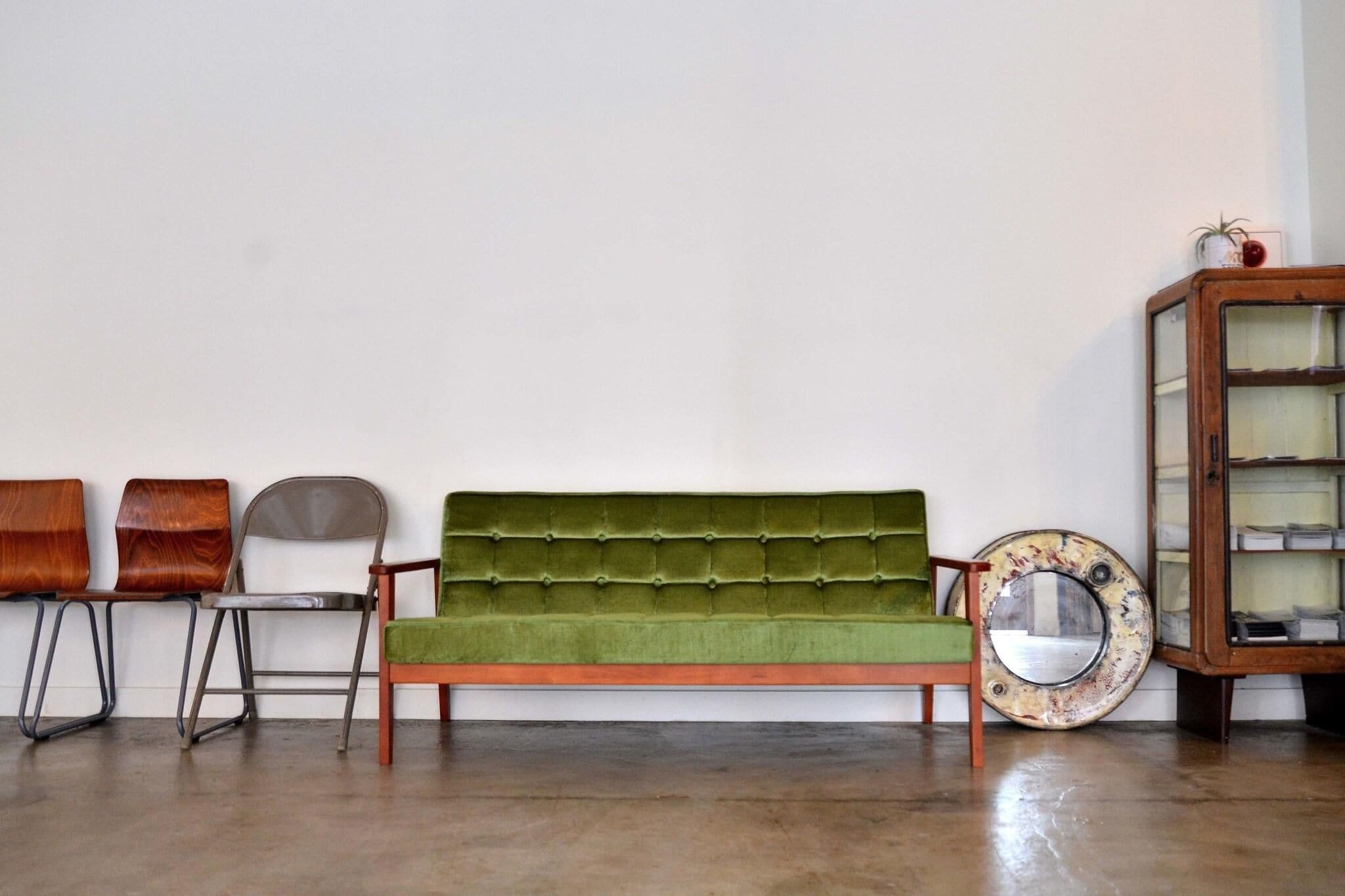 A green sofa, a wooden cabinet, and a chair in a room with a plain wall and a concrete floor.