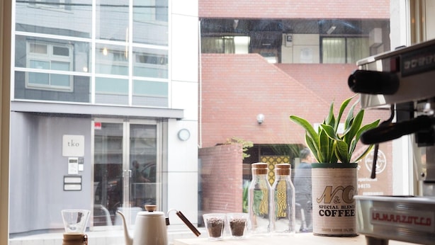 Mostrador de cafetería con una máquina de café, una planta y vista a un edificio desde la ventana.