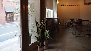 Interior de una cafetería con mesas y sillas, una ventana con plantas y vistas a la calle.