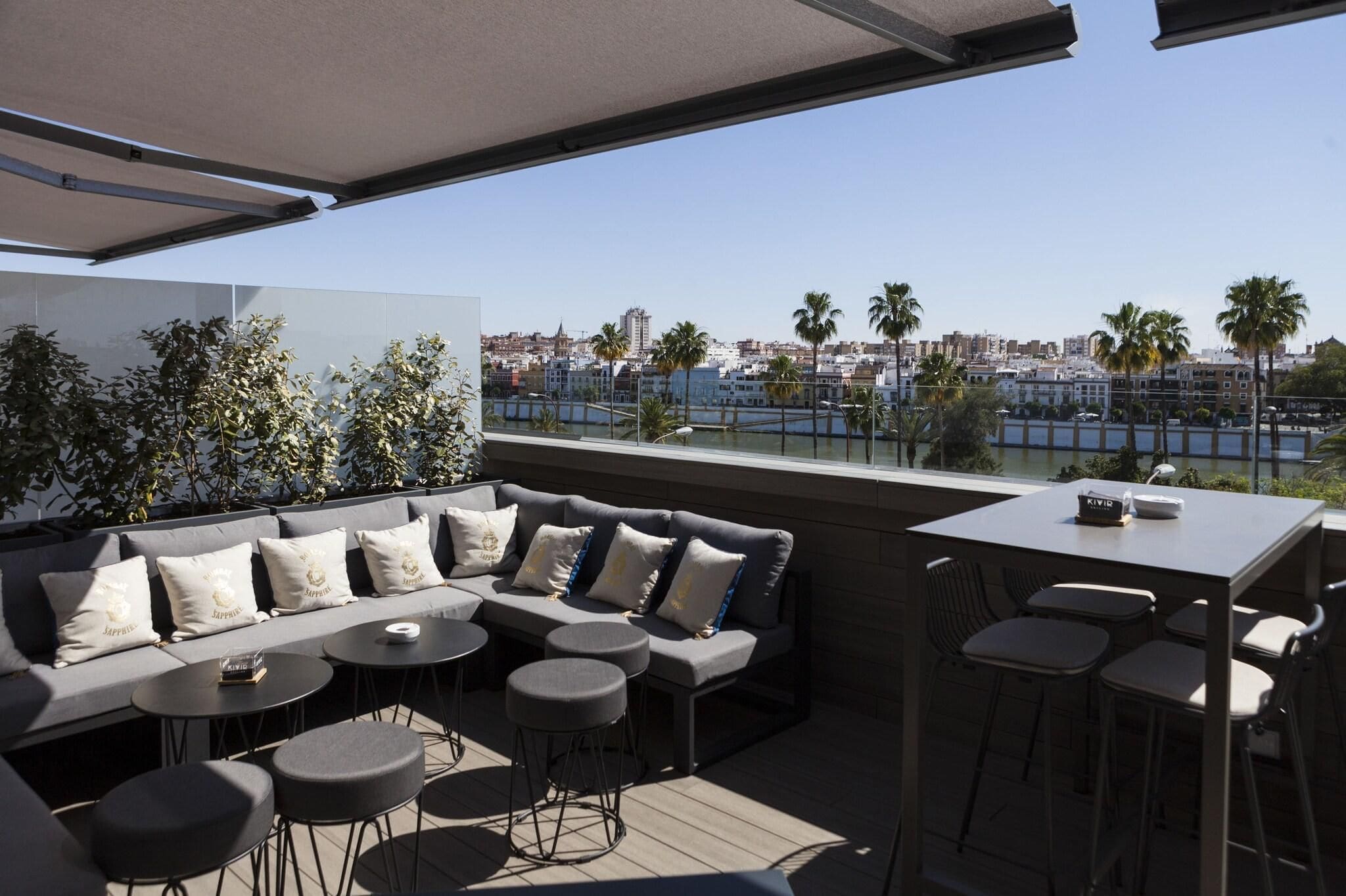 Un espace extérieur aménagé avec un canapé, des tables rondes et des chaises, offrant une vue sur le paysage urbain et le bord de l’eau.