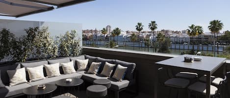 An outdoor seating area with a sofa, round tables, and chairs, overlooking a cityscape and waterfront.