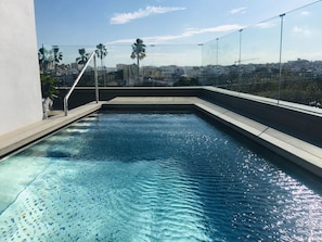 A rooftop pool with clear blue water, a glass barrier, and a cityscape view in the background. - Hotel Kivir Sevilla (Seville)