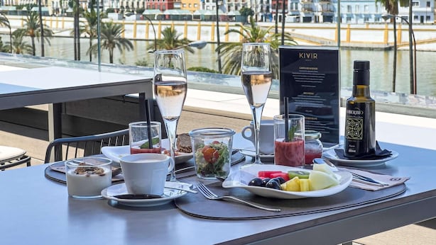 A table set with a glass of water, a cup, a plate of fruit, a bottle of olive oil, and a menu. - Hotel Kivir Sevilla (Seville)