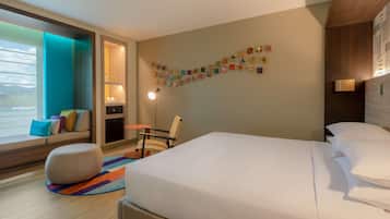A hotel room with a bed, a seating area with cushions, a chair, a small table, a lamp, and a wall with colorful postcards.