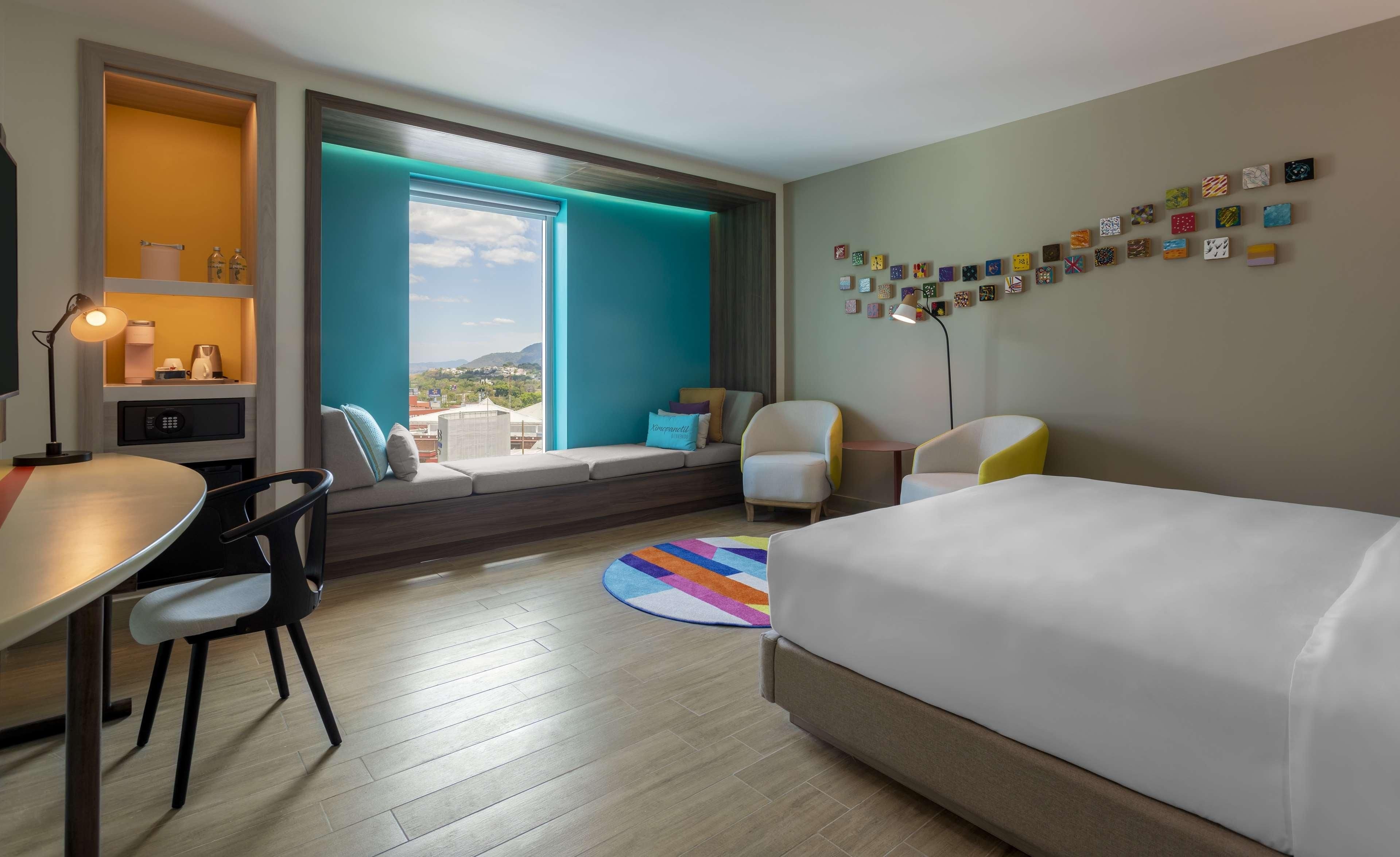 A hotel room with a bed, a desk, a chair, a sofa, a window with a view, and a wall with artwork.
