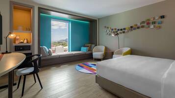 A hotel room with a bed, a desk, a chair, a sofa, a window with a view, and a wall with artwork.