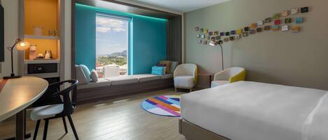 A hotel room with a bed, a desk, a chair, a sofa, a window with a view, and a wall with artwork.