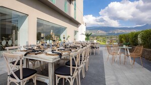 An outdoor dining area with tables and chairs, a building with large windows, and a mountainous landscape in the background.