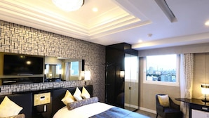 Quarto de hotel com uma cama grande, televisão, secretária com abajur e vista para a cidade.