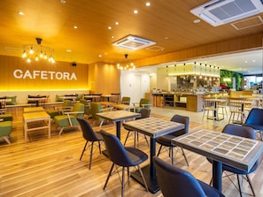 Interior de una cafetería con suelo de madera, sillas verdes y negras, mesas con tops estampados y una zona de cocina al fondo.