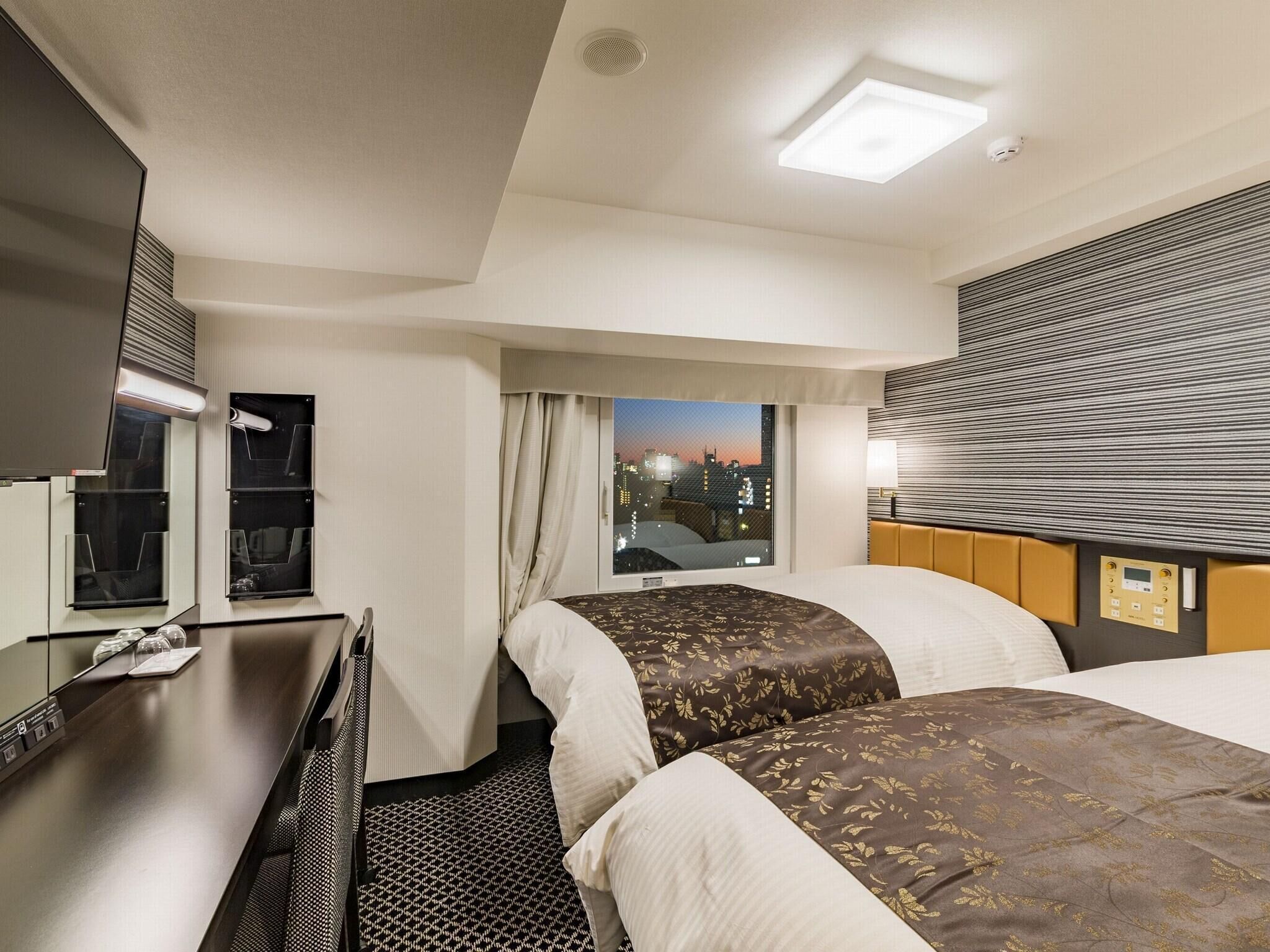 A hotel room with two beds, a desk, a mirror, and a window with a city view.