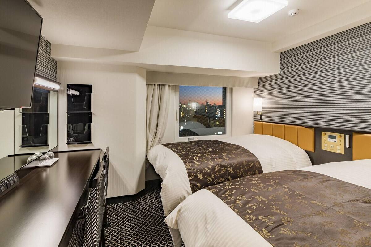 A hotel room with two beds, a desk, a mirror, and a window with a city view.