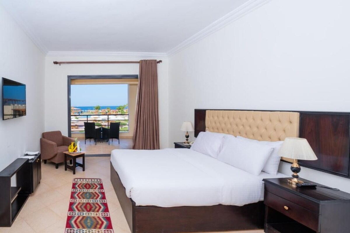 A hotel room with a bed, a desk, a chair, a TV, and a view of the sea.