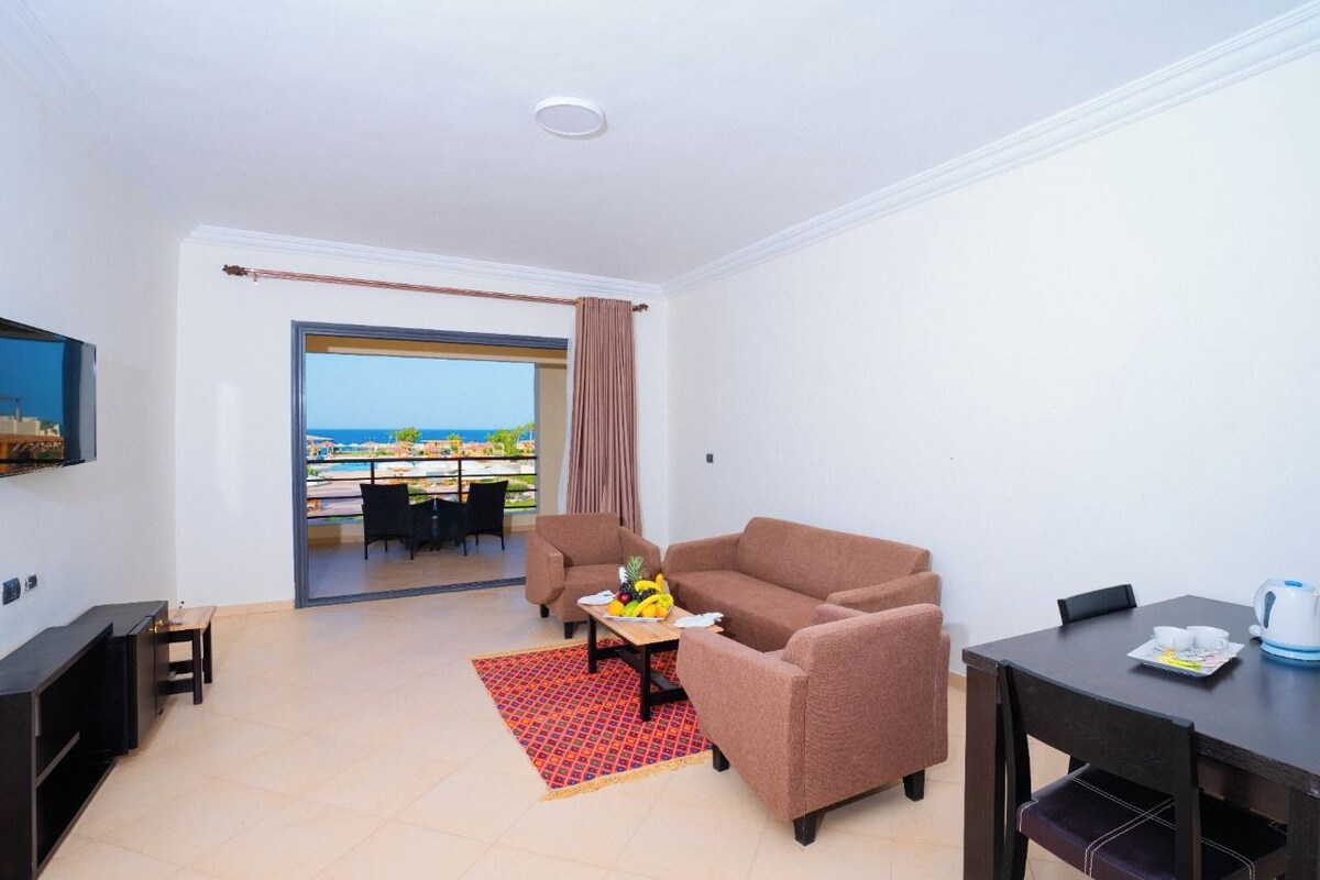 Ein Wohnzimmer mit einer Couch, einem Couchtisch mit Früchten, einem Fernseher und Blick auf das Meer durch ein großes Fenster.